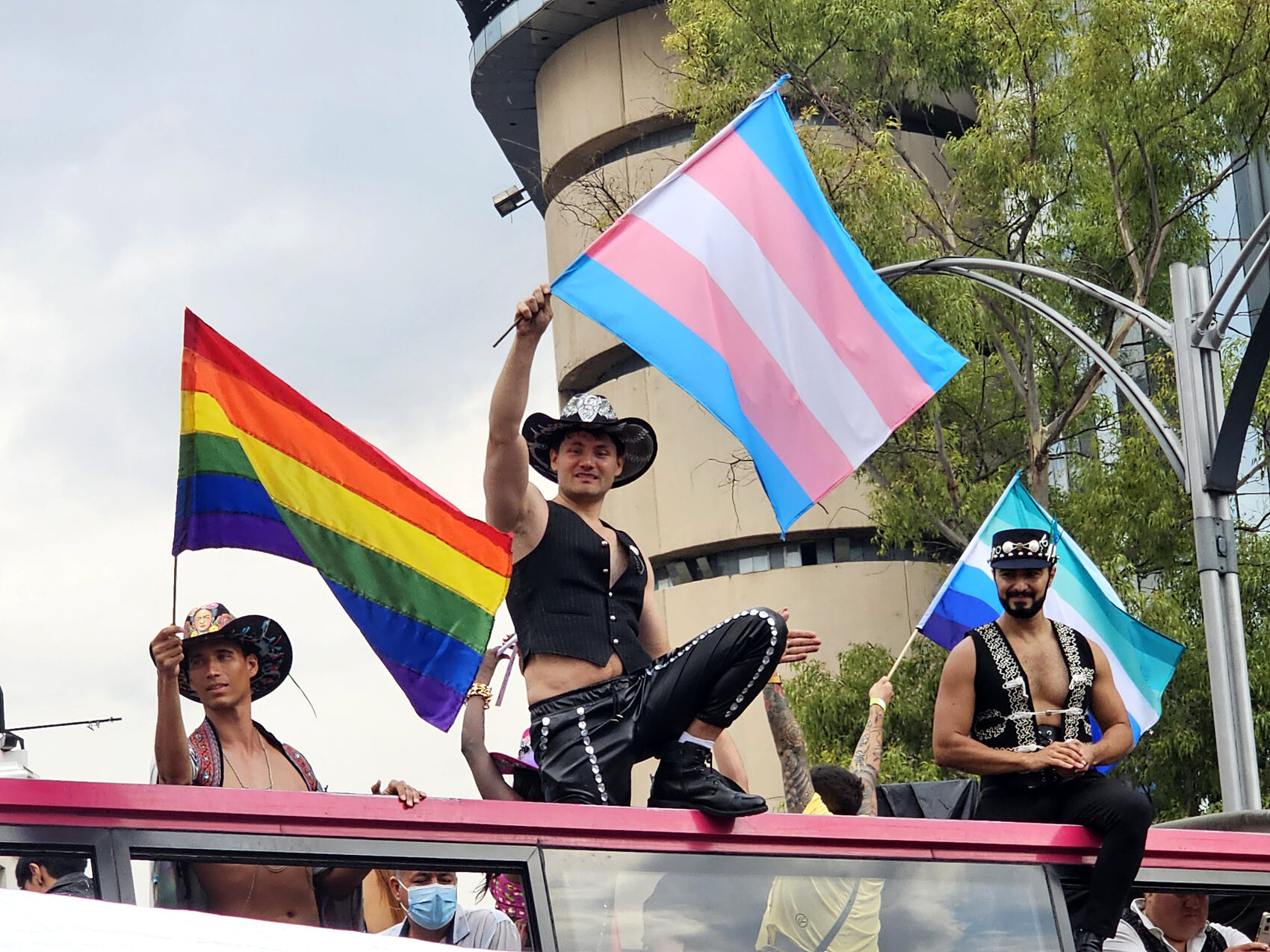 Three men holding trans and gay pride flags on top of a bus in Mexico City