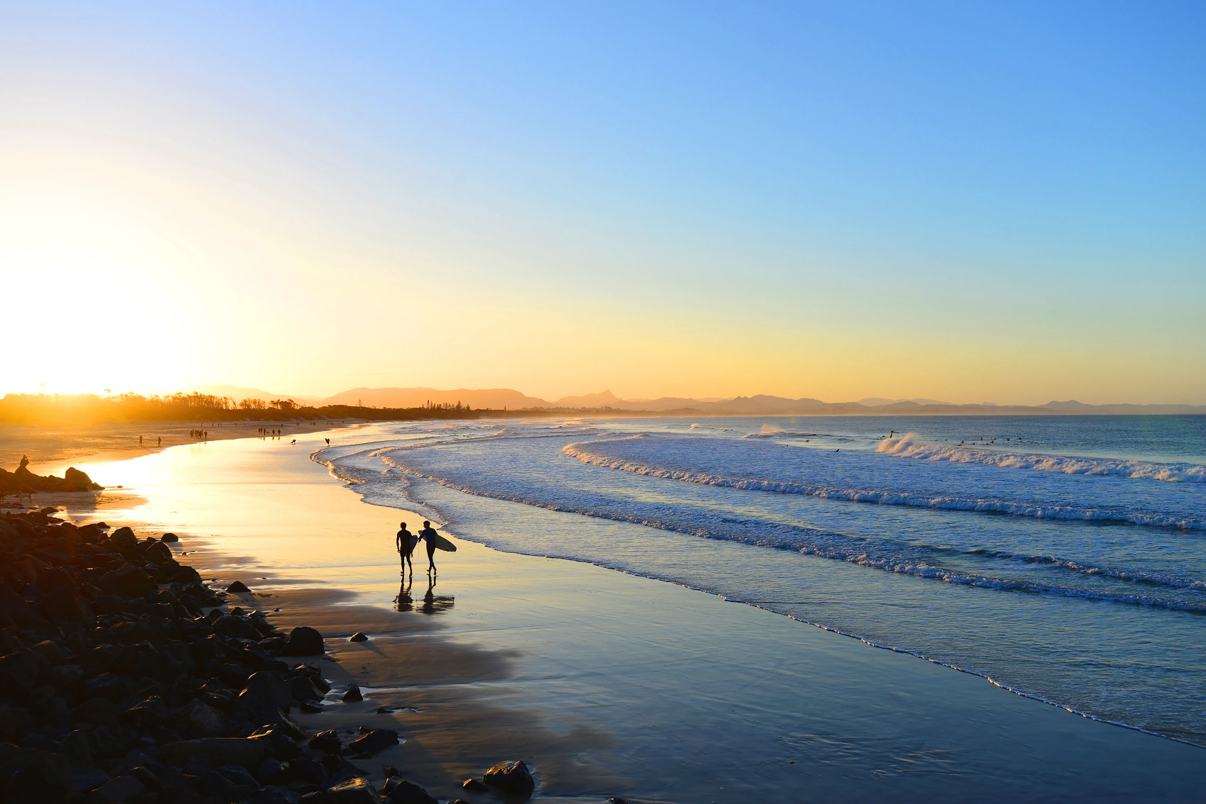 Two surfers walk along the beach at sunset with their surfboards