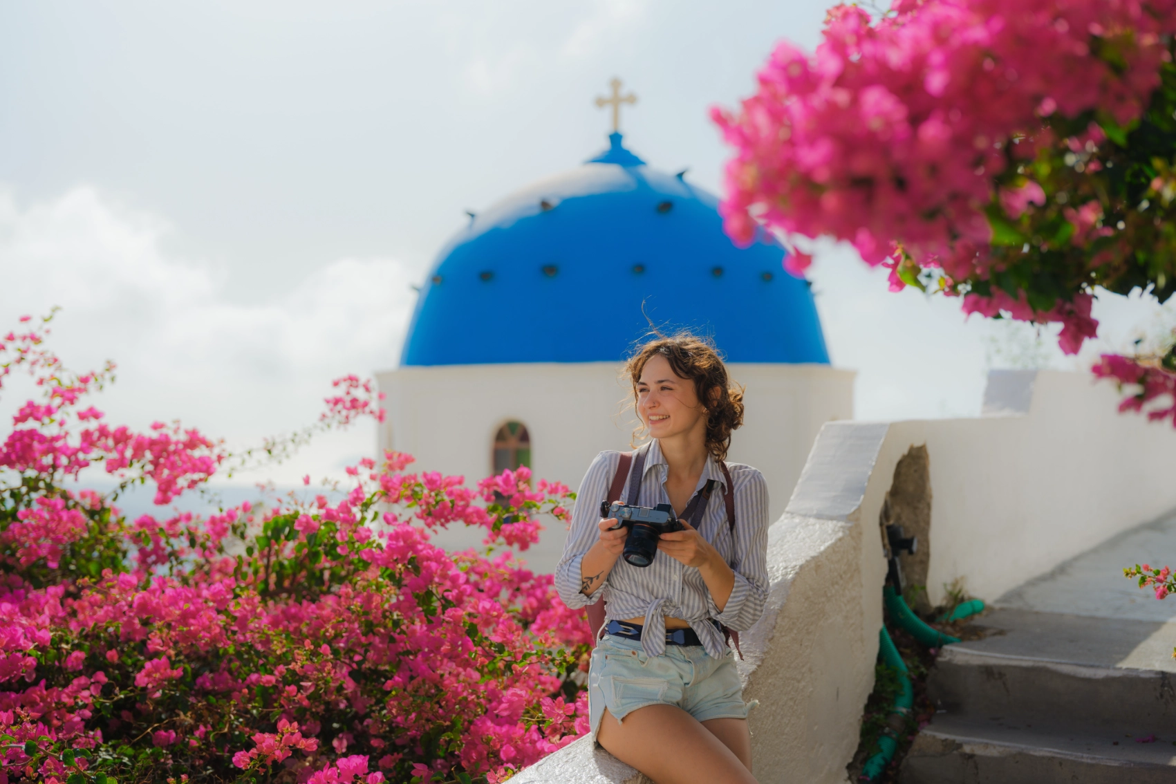 Santorini, Greece A woman traveller sits in front of a whitewashed church with a blue roof surrounded by bright pink flowers in Santorini, Greece