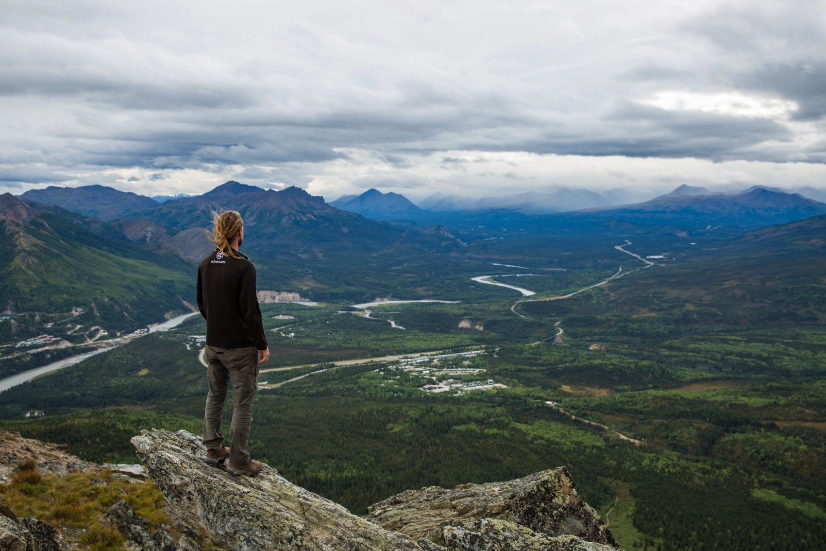 A male traveller stands on a rocky ledge looking out on the landscape on the Mt Healy Overlook Trail in Alaska