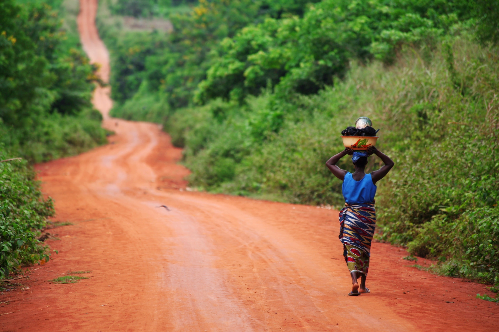 A solo woman walks down a dirt road with a large filled bowl on her head