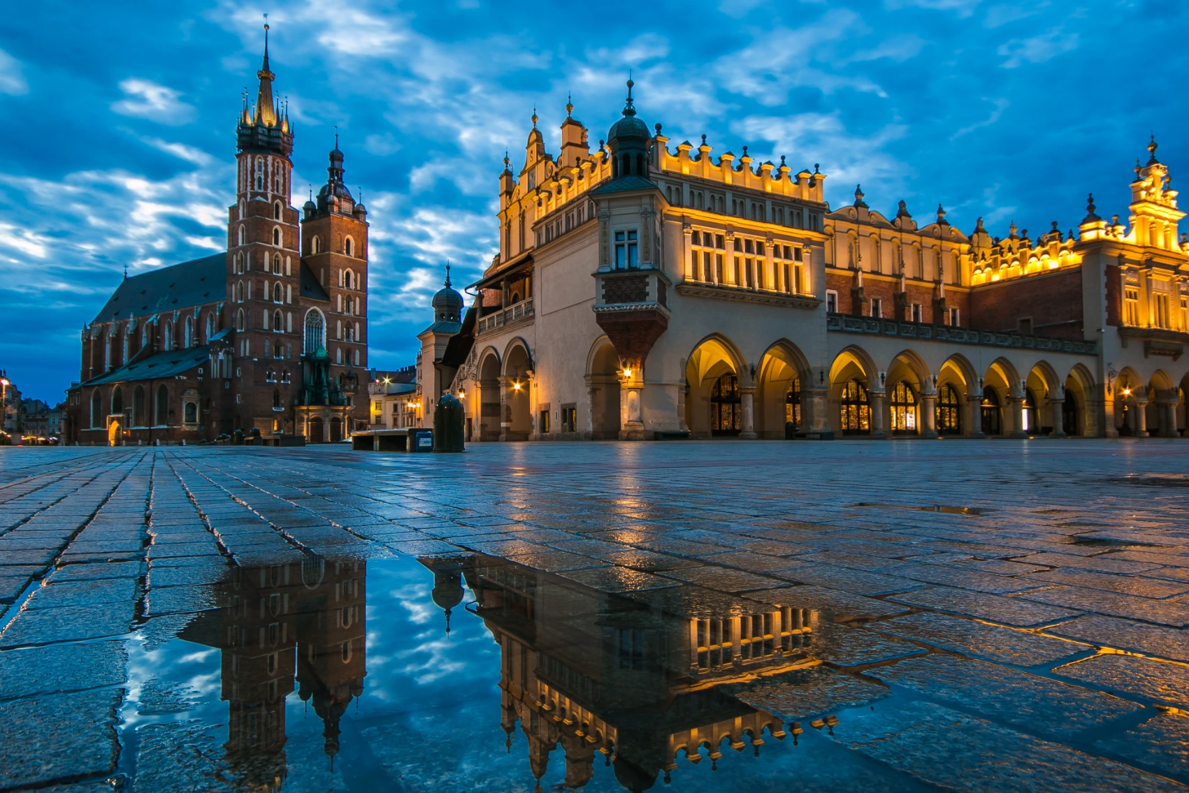 The buildings of Rynek Główny, Kraków's market square, are reflected in puddles of rain at sunrise on a cloudy day in Poland