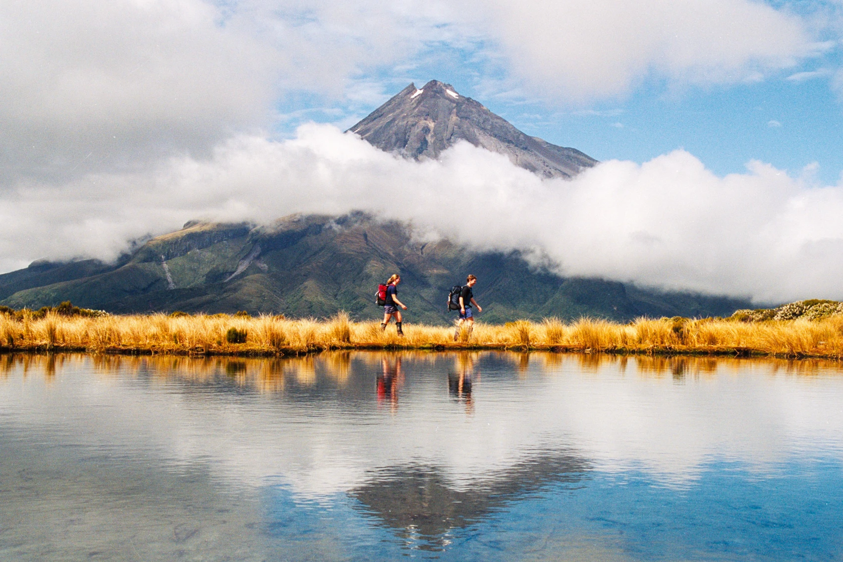 Hikers reflected in a lake walk in front of Mt Taranaki in New Zealand