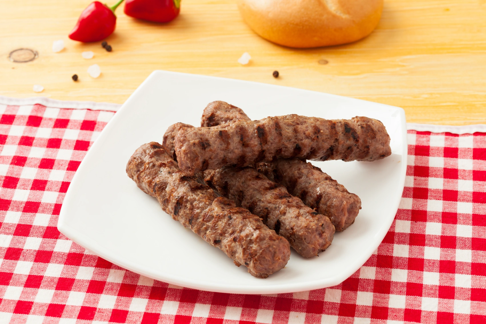 A plate of kebapcheta on a red and white checkered tablecloth with vegetables and bread in the background