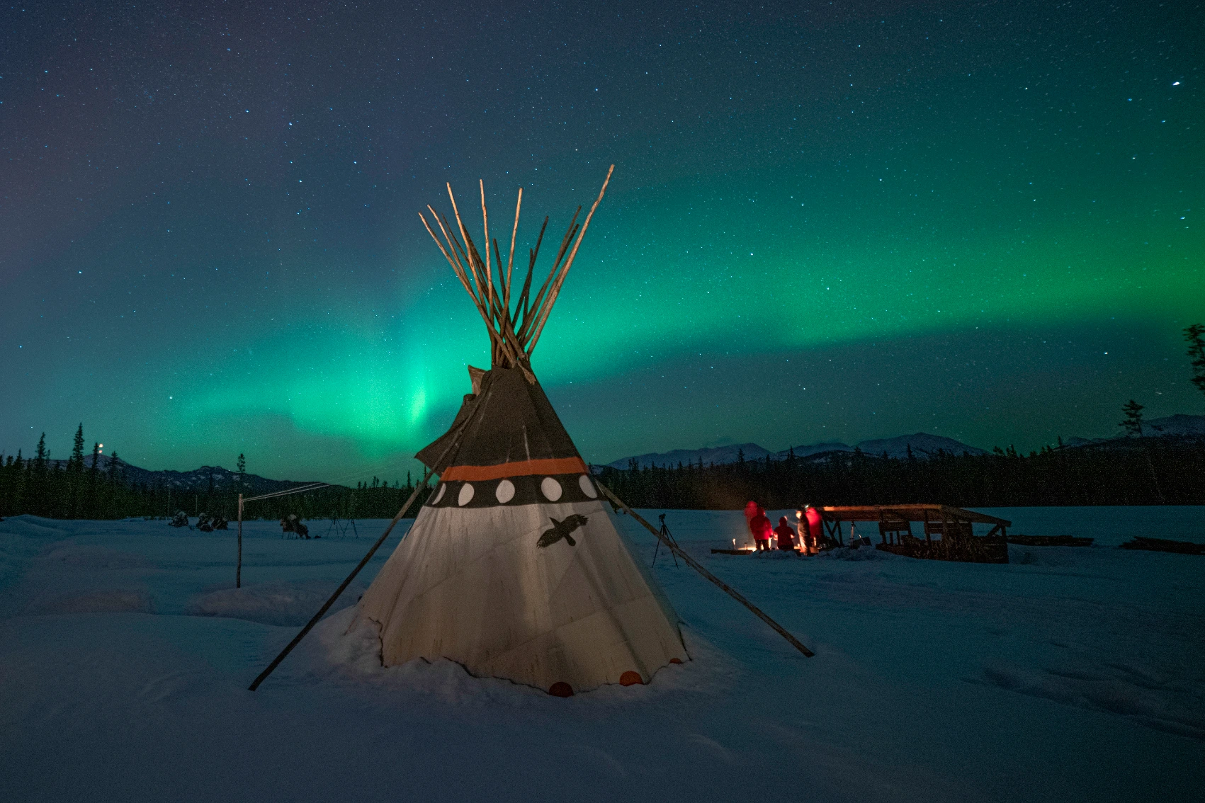 Group of travellers sitting around a campfire next to a teepee under the Northern Lights in the Yukon, Canada
