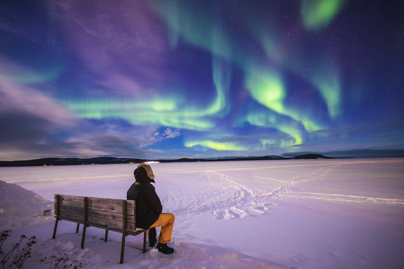 A traveller in a heavy coat sits on a bench and looks out over the snow at the Northern Lights in Finland