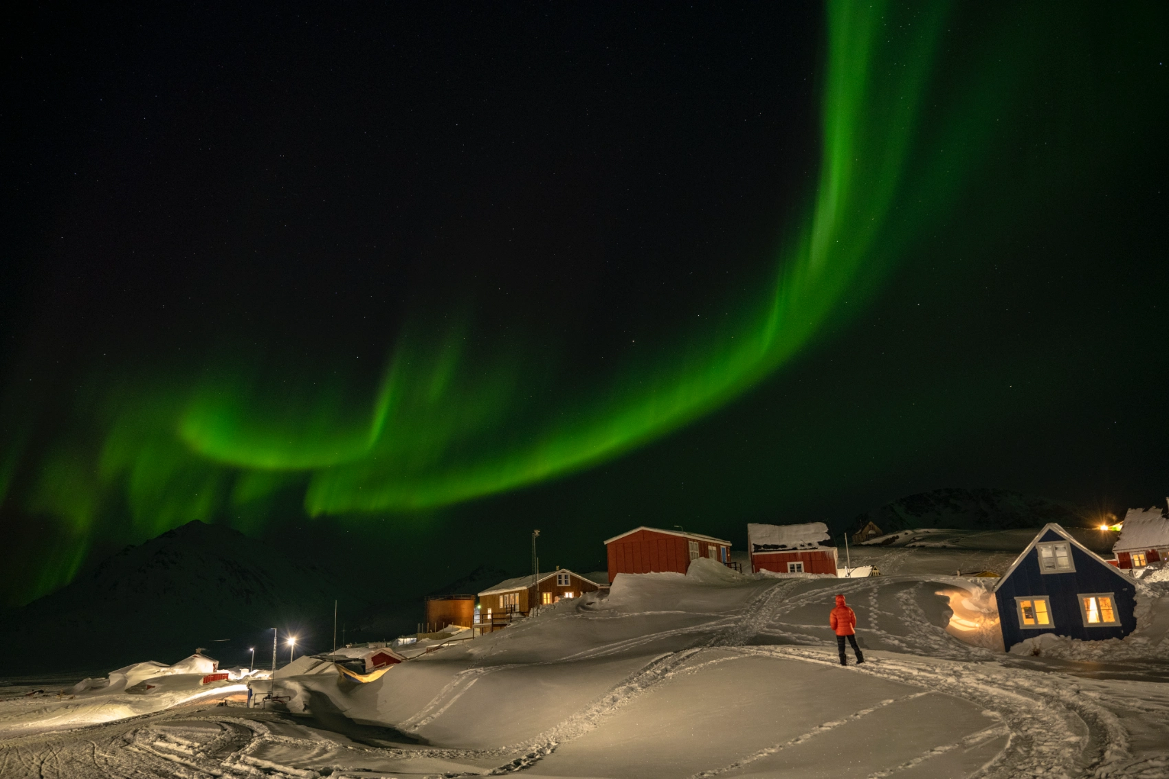 A person in a red coat walks through a small village in Greenland with the Northern Lights overhead