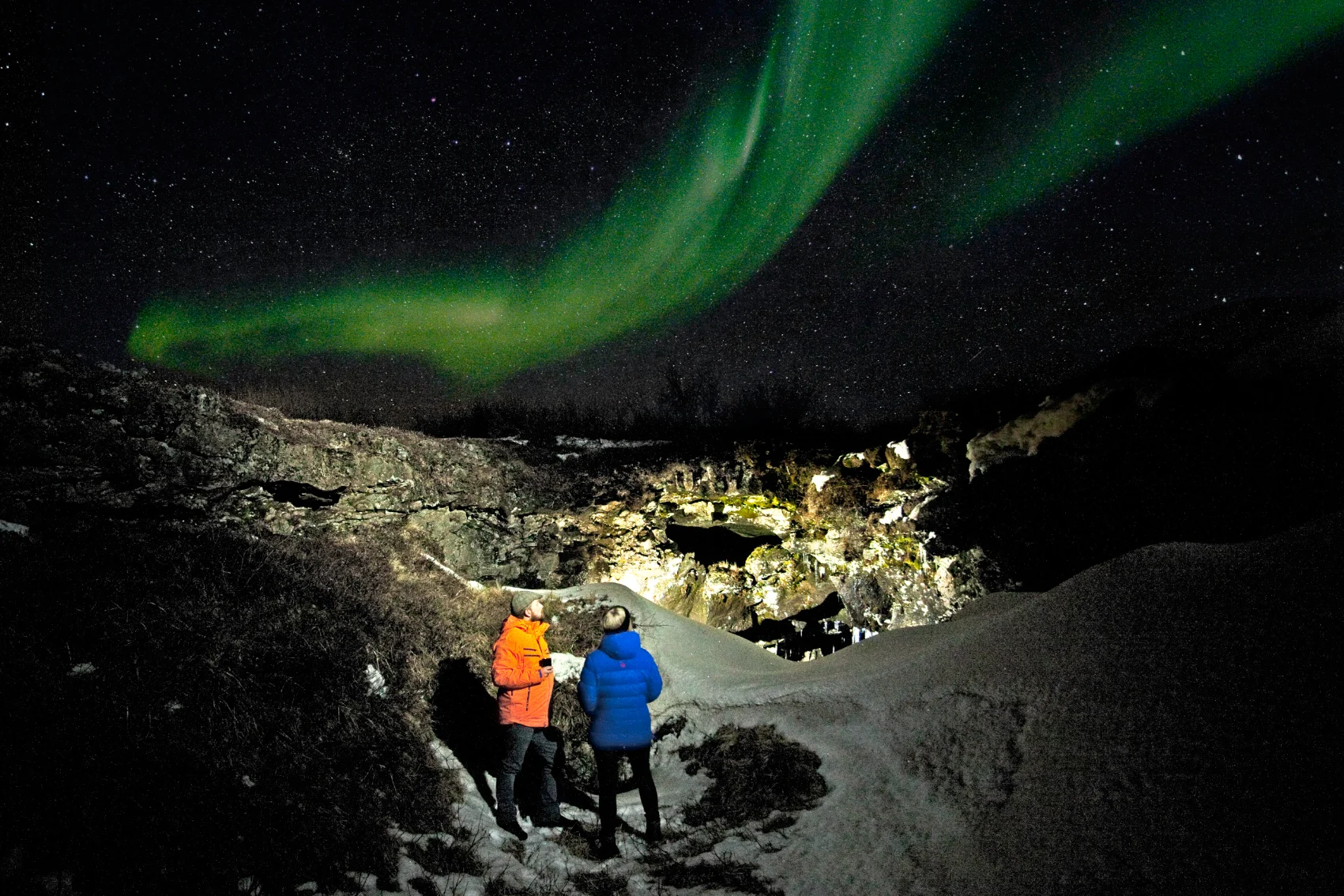 Two travellers stand below a green streak of the Northern Lights in Iceland