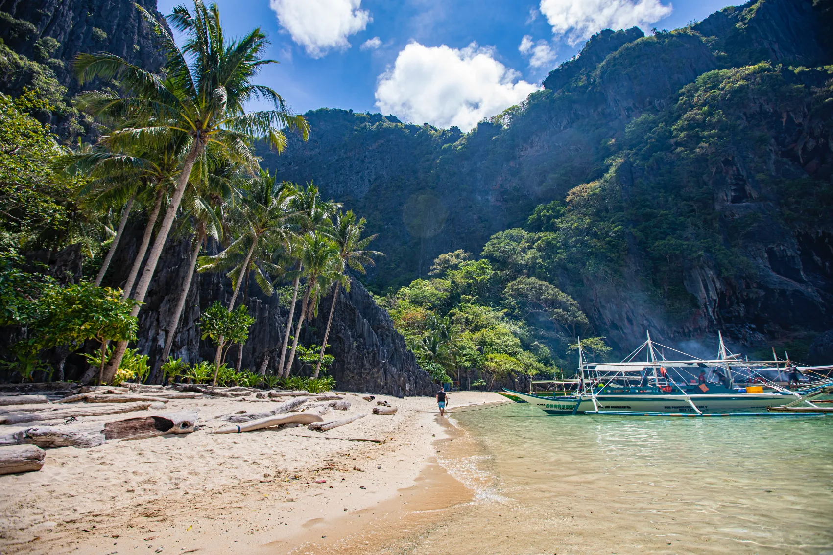 A traveller walks on the sand on El Nido Beach between palm trees and a boat