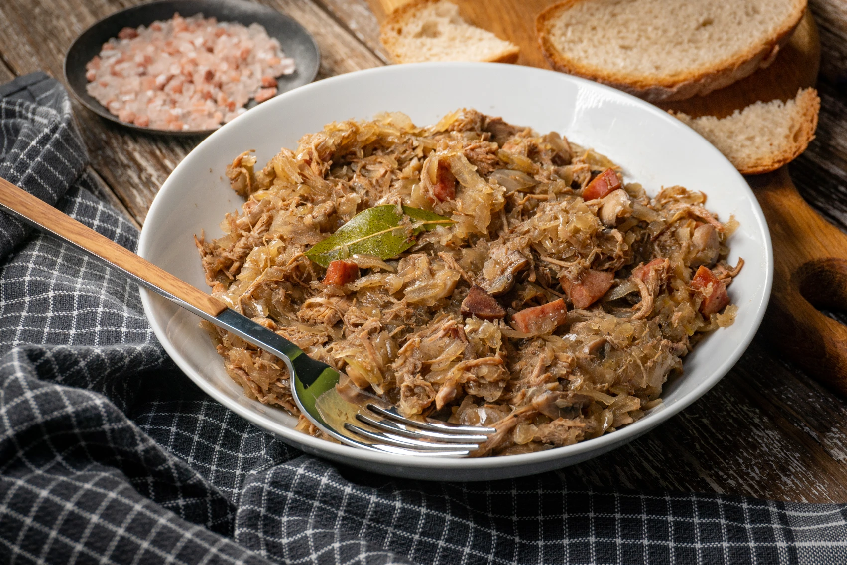A bowl of bigos with bread on the table in the background