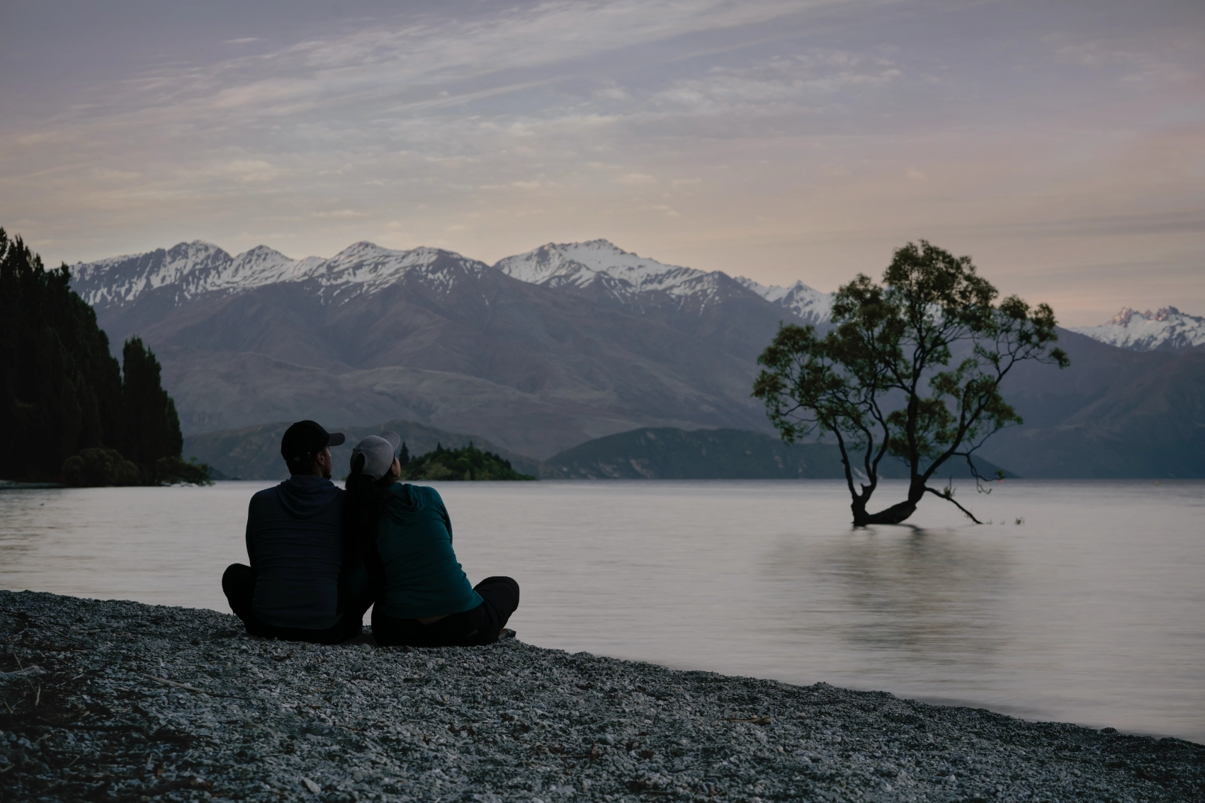 A couple sits in front of the famous That Wanaka Tree at dusk near Queenstown, New Zealand