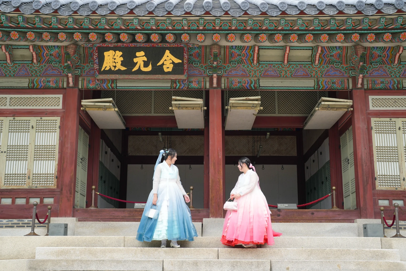 Two Korean women wearing pink and sky-blue hanbok stand in front of the facade of Gyeongbokgung Palace in Seoul, South Korea