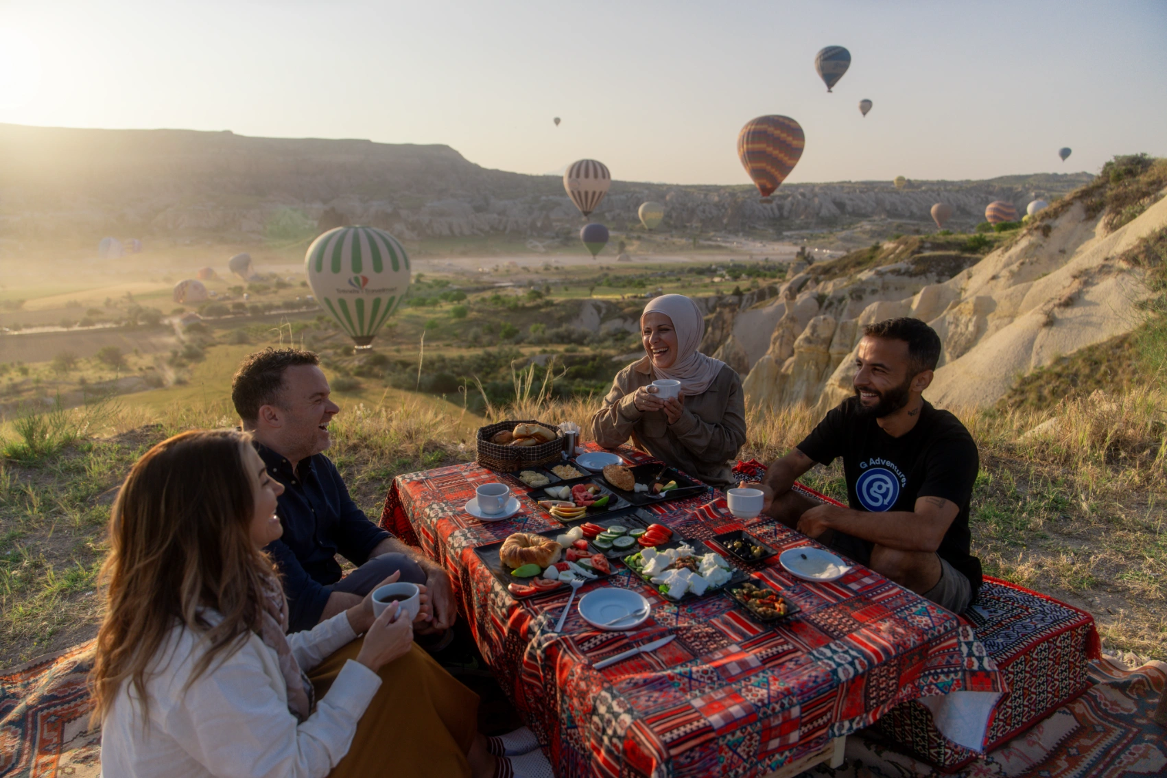 A group of travellers eats breakfast at an outdoor table in Cappadocia, Turkey, with hot-air balloons and fairy chimneys in the background
