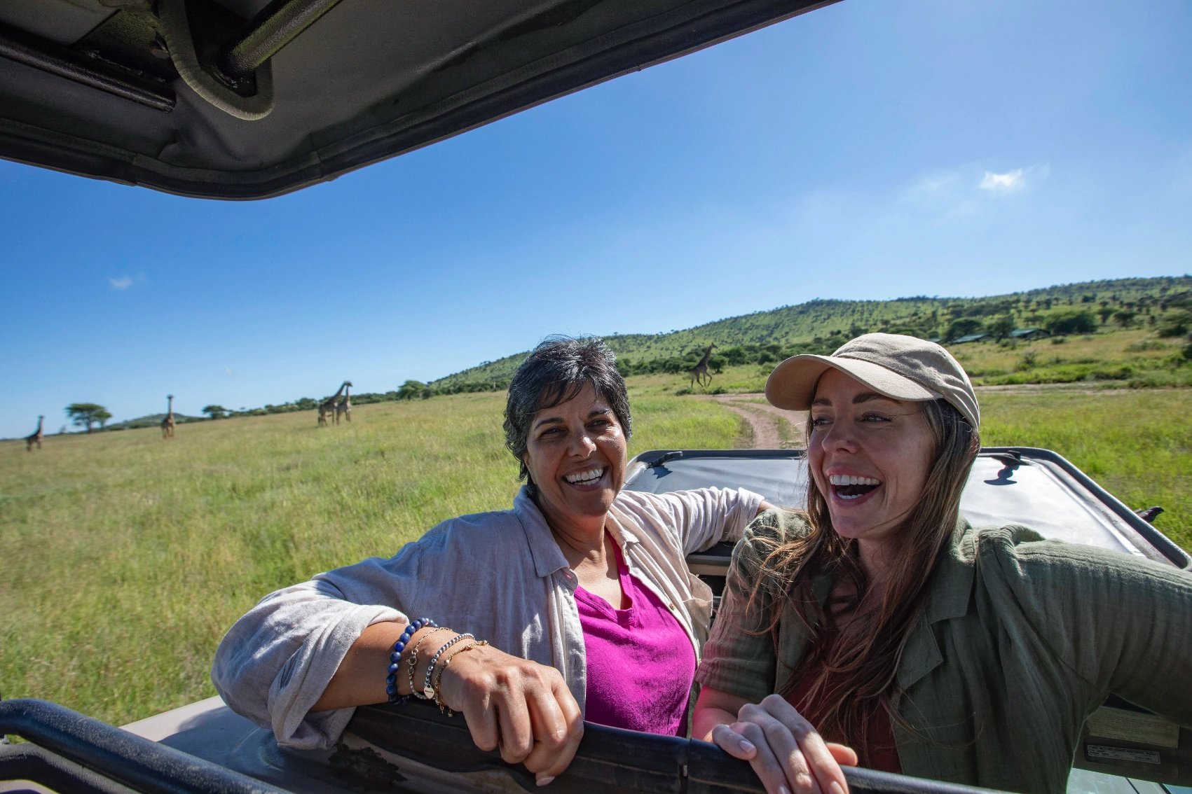 Safari in Tanzania Two female travellers sit in a safari car with giraffes in the background in Tanzania