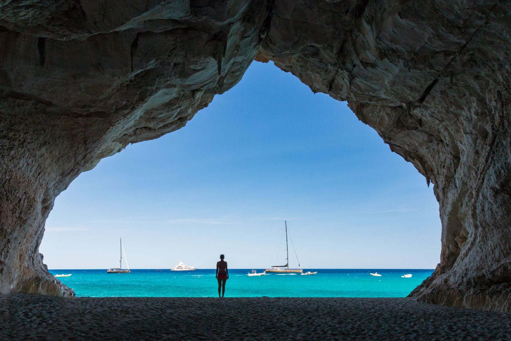 A woman traveller is framed in a rock formation, silhouetted by the blue ocean
