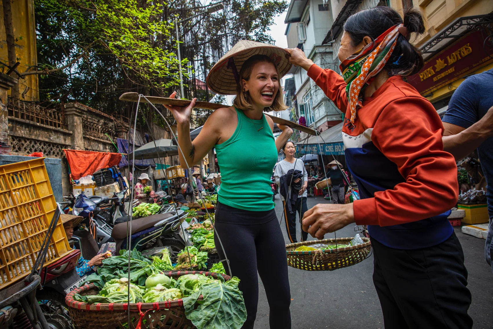 A Vietnamese woman puts a hat on a female traveller holding baskets of vegetables at a market in Vietnam