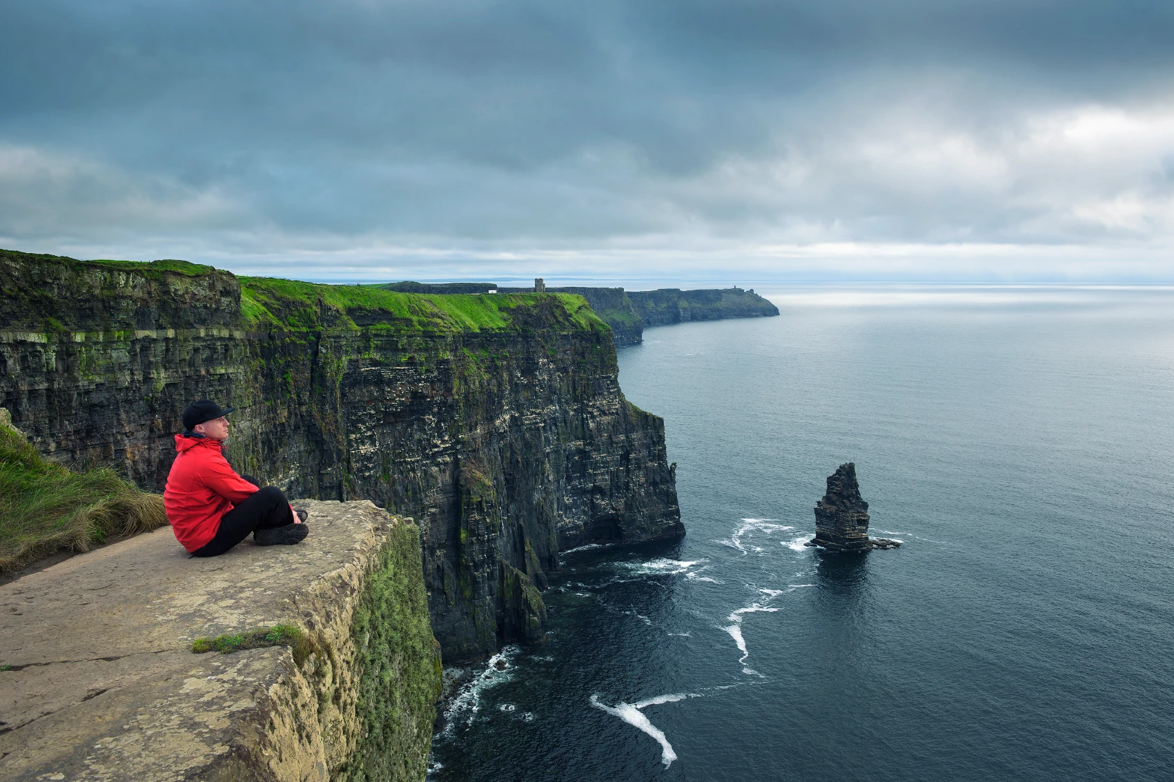 A hiker sits on a rock ledge overlooking the Atlantic Ocean and the Cliffs of Moher in Ireland