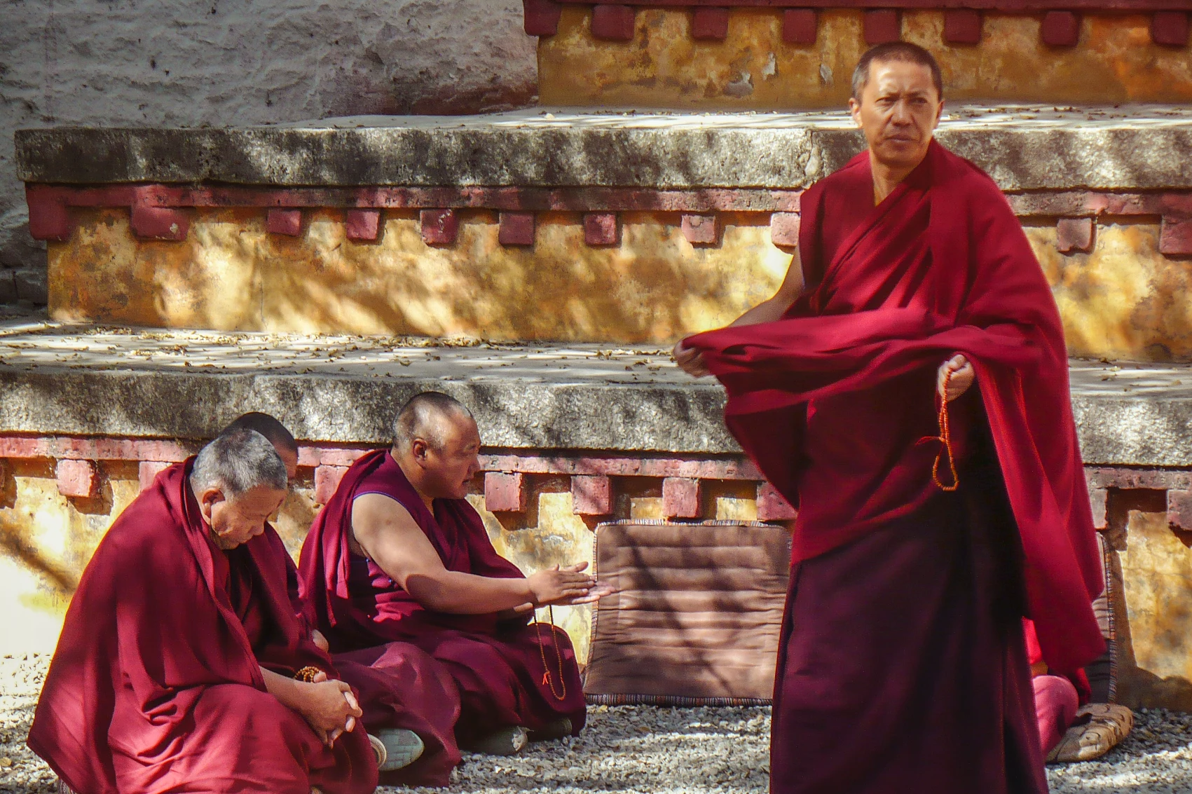A monk in maroon robes walk past two seated monks in Lhasa, Tibet