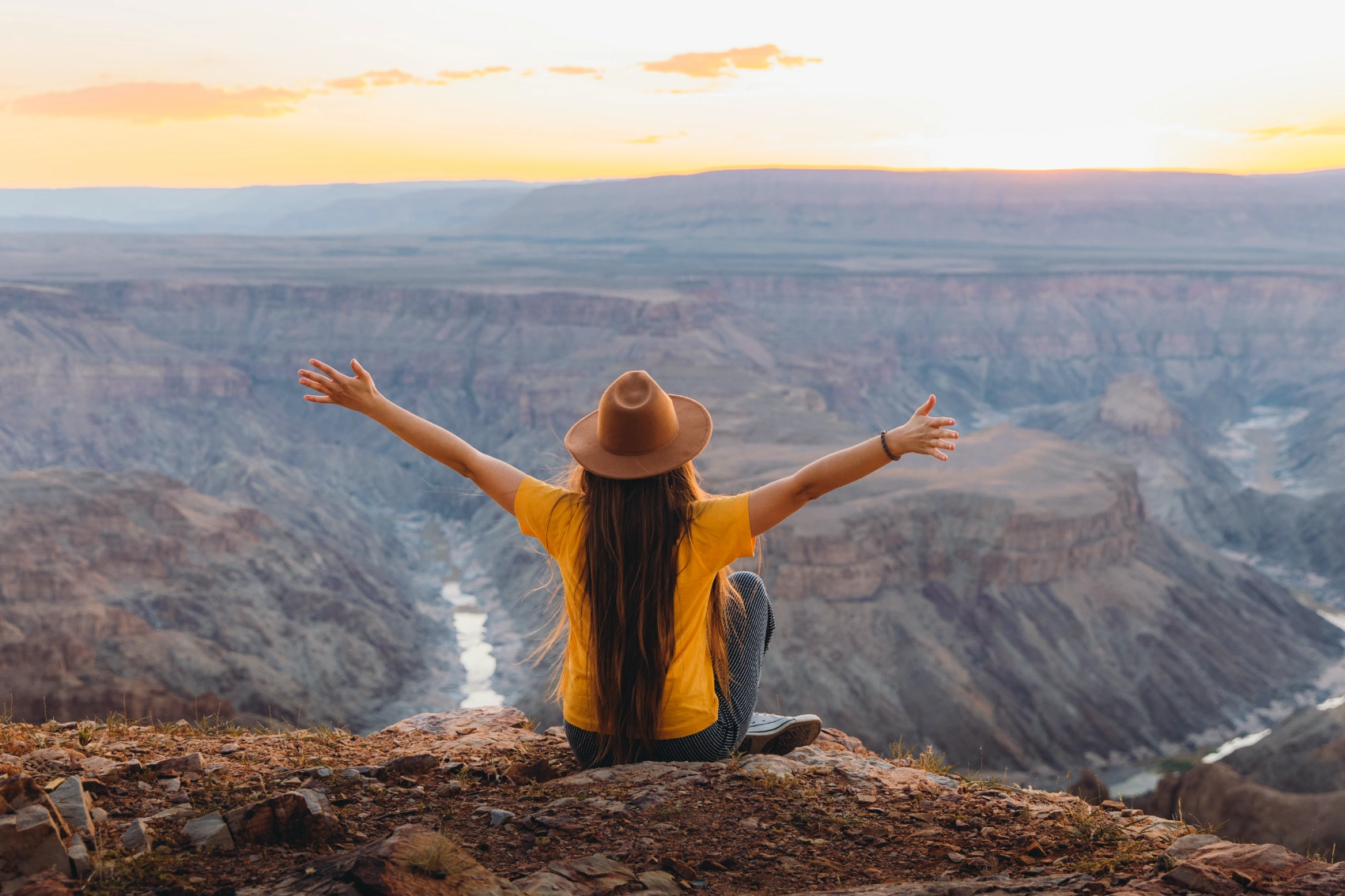 A woman traveller sits on the edge of Fish River Canyon, Namibia, while raising her arms at sunset