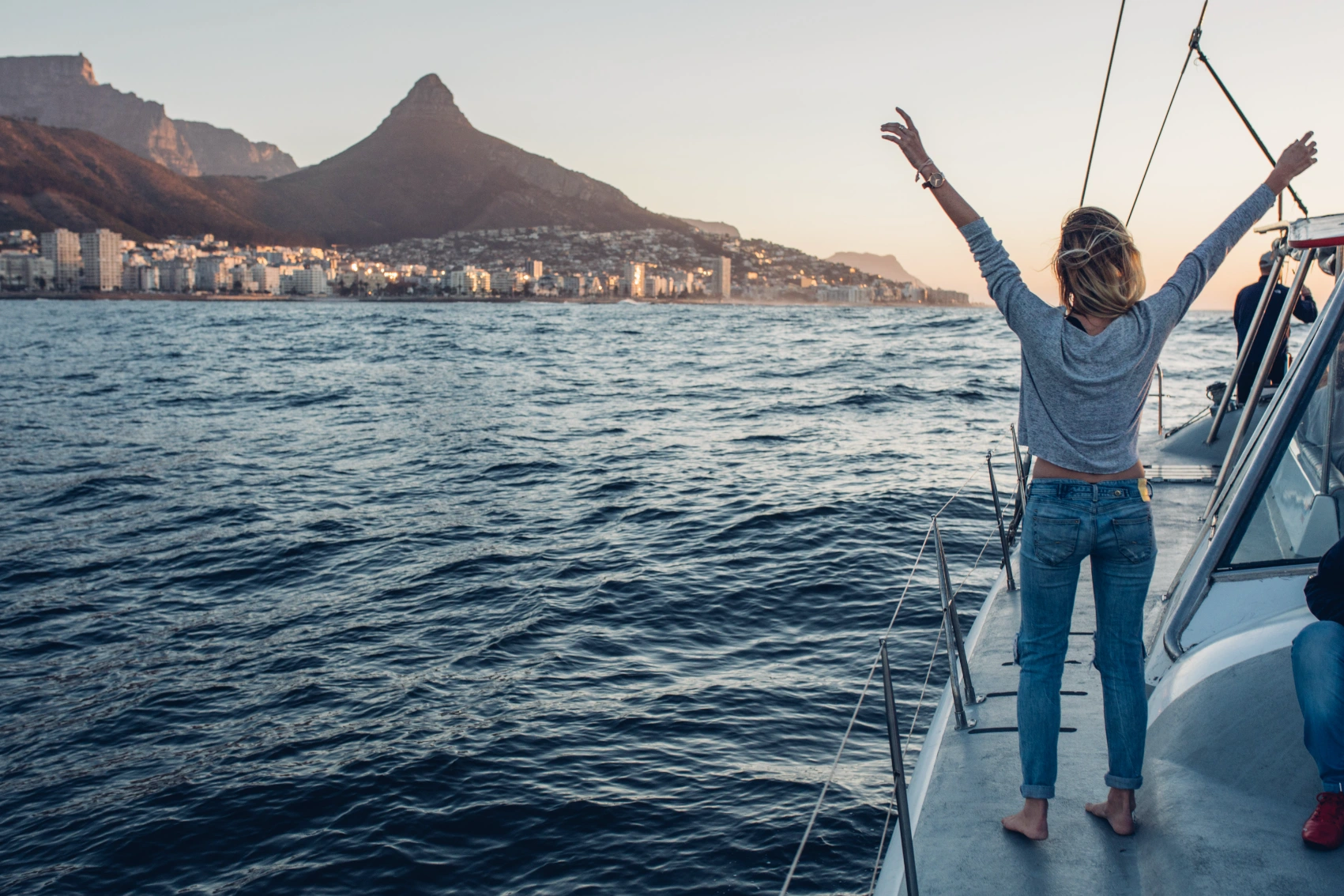 A woman traveller on a boat raises her arms while looking at the cityscape of Cape Town, South Africa