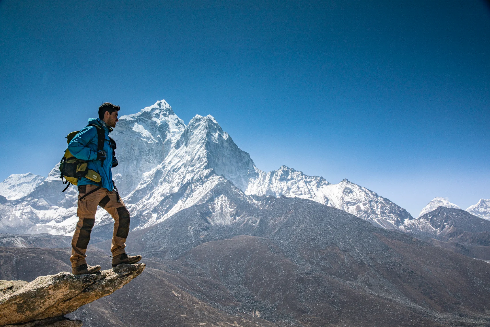A man stands on a rocky ledge in front of a snowy mountain in Nepal
